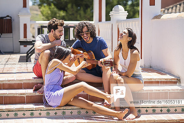 Cheerful young friends enjoying while sitting with guitar on steps at terrace during sunny day