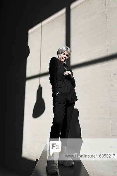Stilvolle Frau in elegantem Anzug steht auf Stützmauer mit Sonnenlicht und Schatten im Hintergrund im Büro