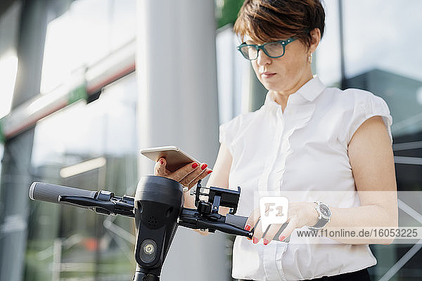 Businesswoman using smart phone while standing with push scooter in city