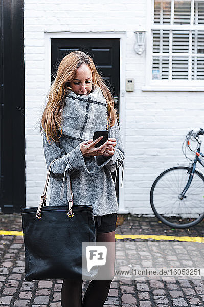 Mittlere erwachsene Frau in warmer Kleidung  die auf einer Straße in der Stadt ein Smartphone benutzt