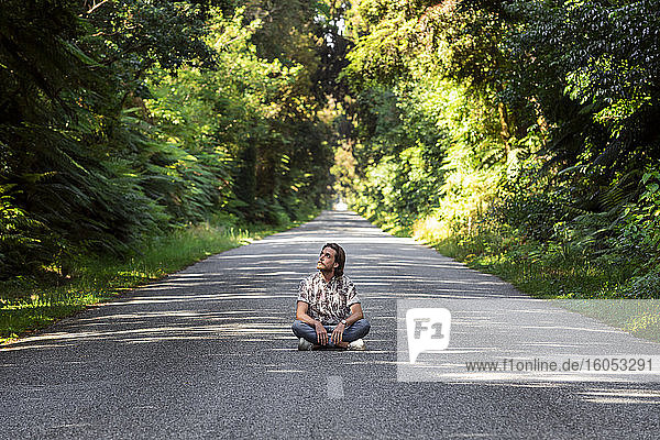 Thoughtful young man sitting on road amidst trees in forest