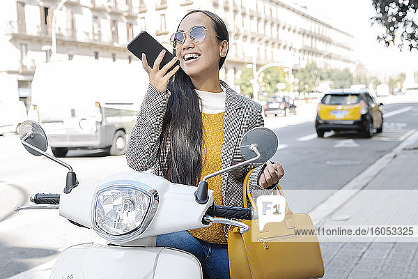 Fröhliche junge Frau  die über ihr Smartphone spricht  während sie auf einem Motorroller sitzt