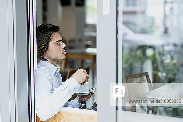 Nachdenklicher Geschäftsmann  der eine Kaffeetasse hält  während er in einem Café sitzt  gesehen durch ein Fenster