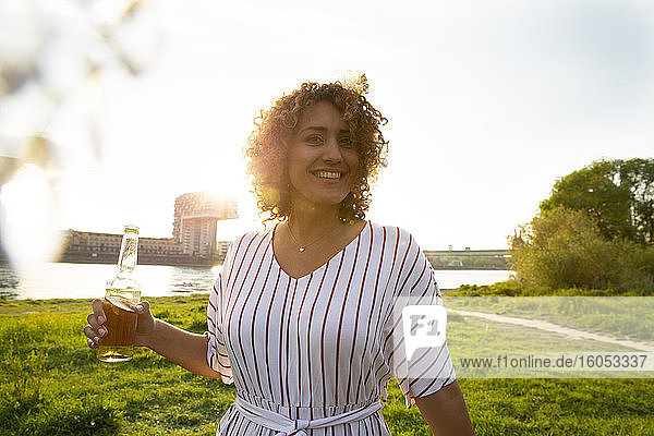 Smiling woman with curly hair holding beer bottle while standing on land against sky