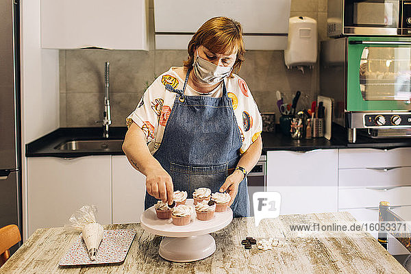 Weibliche Bäckerin mit Maske beim Verzieren von Törtchen auf einem Tortenständer in einer Werkstatt