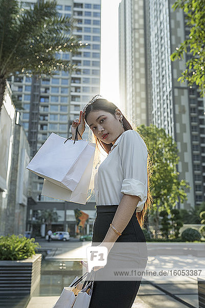 Selbstbewusste Frau  die Einkaufstüten hält  während sie vor modernen Gebäuden in der Stadt steht