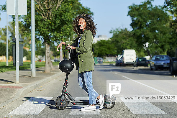 Selbstbewusste junge Frau steht mit elektrischem Roller auf der Straße in der Stadt