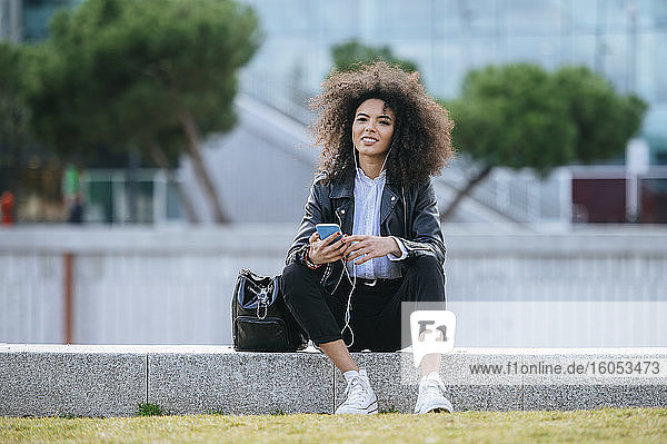 Lächelnde junge Afro-Frau sitzt mit Smartphone und Handtasche auf einer Stützmauer