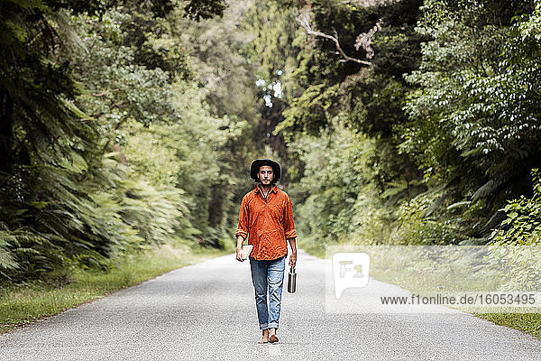 Selbstbewusster junger Mann  der eine Wasserflasche hält  während er auf einer Straße im Wald spazieren geht