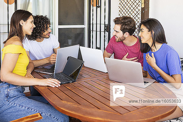 Glückliche junge Männer und Frauen unterhalten sich  während sie mit Laptops am Tisch im Hinterhof sitzen