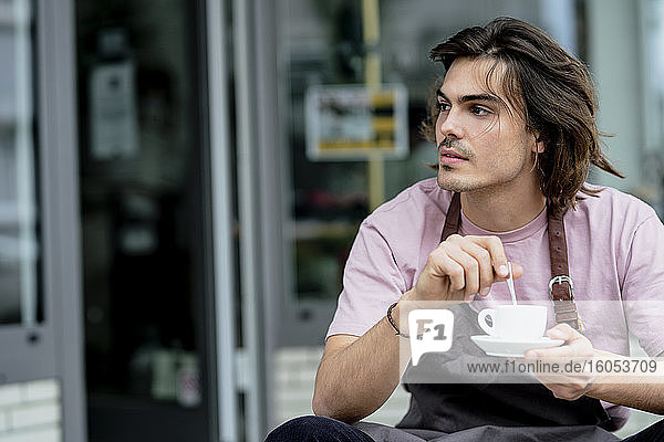 Nachdenklicher männlicher Besitzer hält Kaffeetasse  während er vor einem Café sitzt