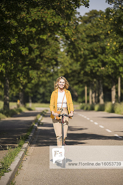 Lächelnde reife Frau fährt elektrischen Roller auf der Straße während des sonnigen Tages
