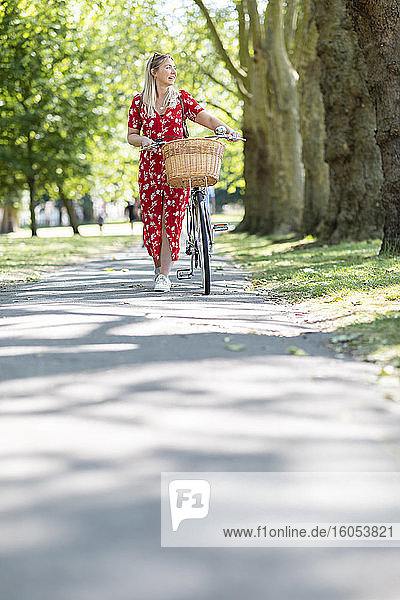 Woman looking away while walking with bicycle on footpath in public park during sunny day