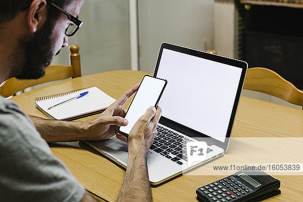 Mittelgroßer erwachsener Mann mit Smartphone und Laptop bei der Arbeit zu Hause