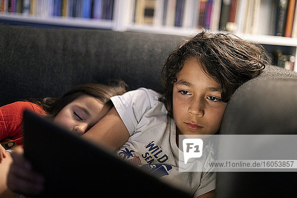 Mädchen schläft neben ihrem Bruder  der ein digitales Tablet benutzt  während er sich zu Hause auf dem Sofa entspannt