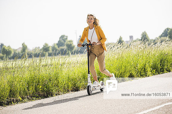 Playful reife Frau Reiten elektrische Push-Roller auf der Straße im Sommer
