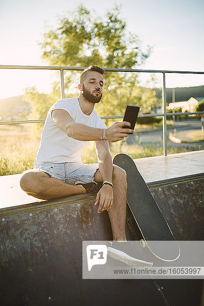 Gut aussehender Mann  der ein Selfie mit seinem Smartphone macht  während er auf einer Sportrampe im Skateboardpark sitzt