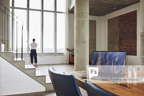 Laptop auf Holztisch in einer Loftwohnung mit Frau am Fenster im Hintergrund