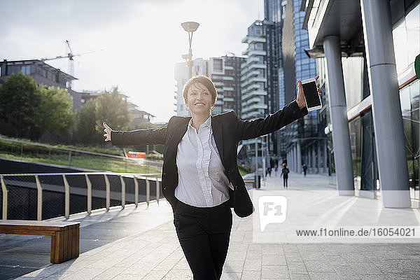 Glückliche Geschäftsfrau mit ausgestreckten Armen  die ein Smartphone hält  während sie auf einem Fußweg in der Stadt spazieren geht