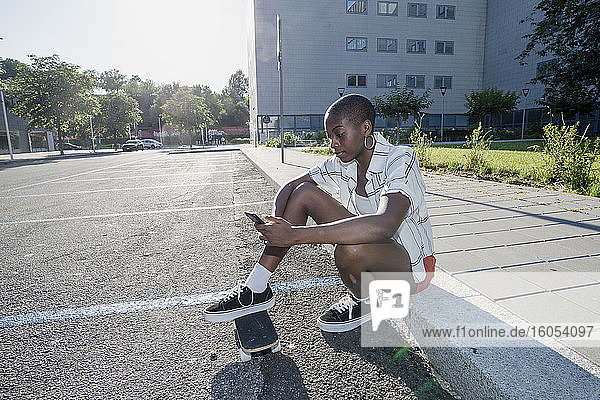 Junge Frau  die ihr Smartphone benutzt  während sie mit ihrem Skateboard auf einem Fußweg in der Stadt sitzt