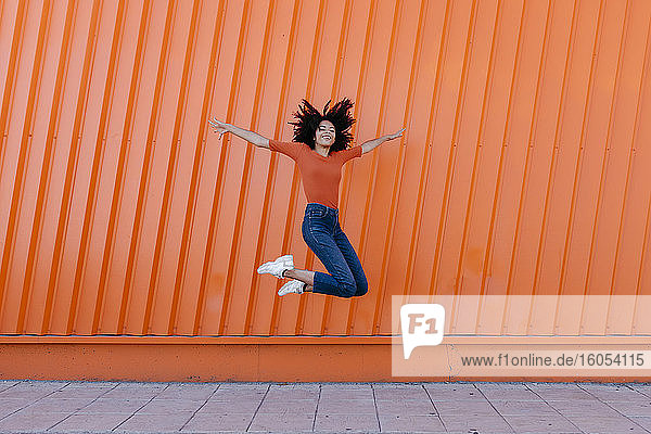 Glückliche junge Frau  die mit ausgestreckten Armen gegen eine orangefarbene Wand springt