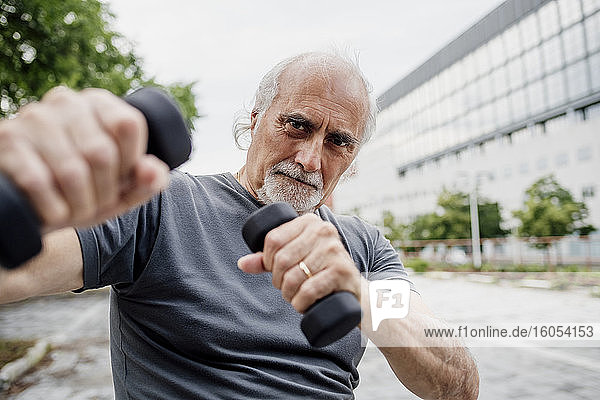 Close-up of confident senior man holding dumbbells while standing in city