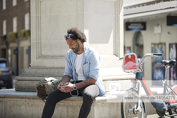 Young man with rental bike listening music with cell phone and earphones in the city  London  UK