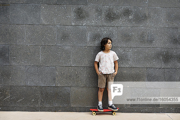 Nachdenklicher Junge mit Händen in den Taschen  der auf einem Skateboard an der Wand steht
