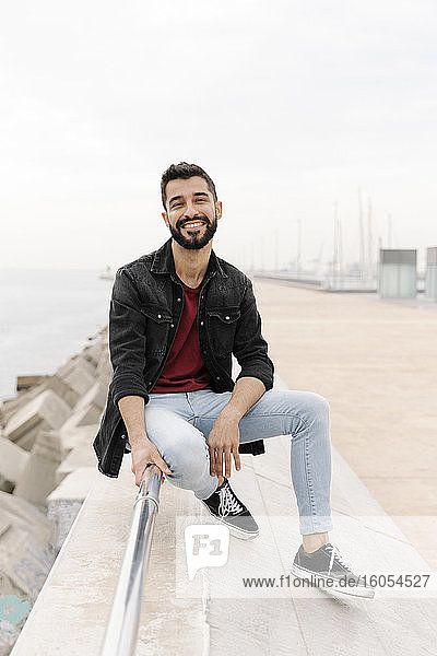 Smiling young man sitting on railing against clear sky in city