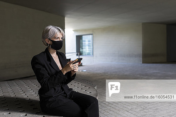 Businesswoman wearing mask using smart phone while sitting on seat in building