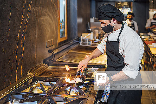 Chef wearing protective face mask preparing a dish in frying pan in restaurant kitchen