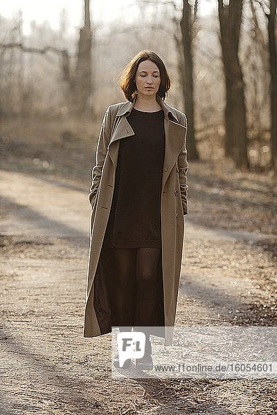 Mature woman with hands in overcoat pockets walking on road at forest