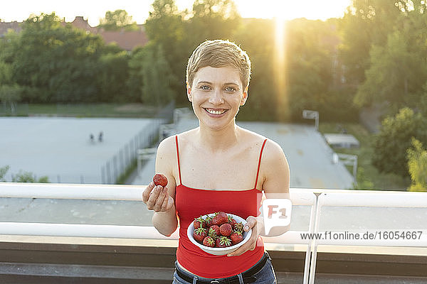 Lächelnde schöne Frau  die Erdbeeren isst  während sie bei Sonnenuntergang auf dem Balkon steht
