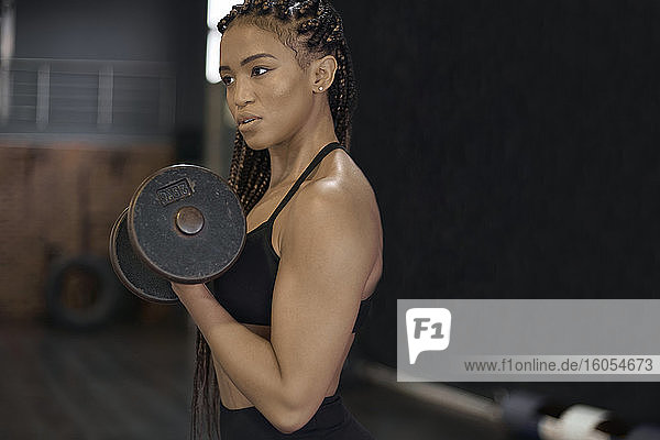 Confident female athlete lifting dumbbells looking away while standing in gym