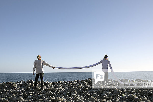 Frau mit langen künstlichen Armen hält die Hand eines Mannes auf Kieselsteinen am Strand gegen den klaren Himmel