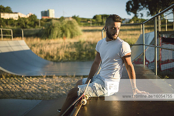 Nachdenklicher junger Mann  der wegschaut  während er auf einer Sportrampe im Skateboardpark sitzt