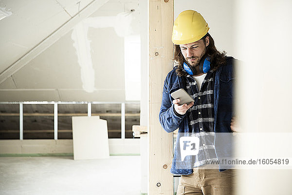 Bauarbeiter mit Helm  der sein Smartphone benutzt  während er bei der Renovierung eines Hauses am Holz steht