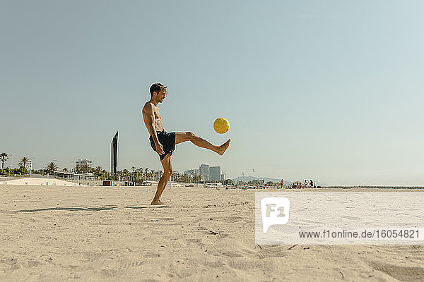 Hemdloser junger Mann spielt mit Ball am Strand gegen klaren Himmel an einem sonnigen Tag