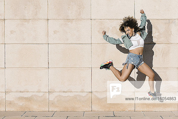 Excited young woman jumping against wall during sunny day