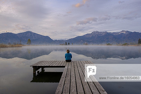 Deutschland  Bayern  Schlehdorf  Frau sitzt am Ende des Stegs und bewundert den Eichsee bei nebligem Morgengrauen