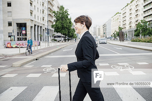 Smiling businesswoman with suitcase crossing road in city