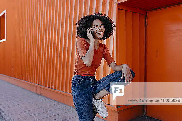 Fröhliche junge Frau  die mit ihrem Smartphone vor einer orangefarbenen Struktur spricht
