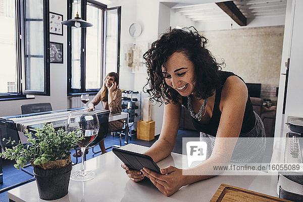 Lächelnde junge Frau  die ein digitales Tablet benutzt  während sie sich bei einem Freund zu Hause auf den Tisch lehnt
