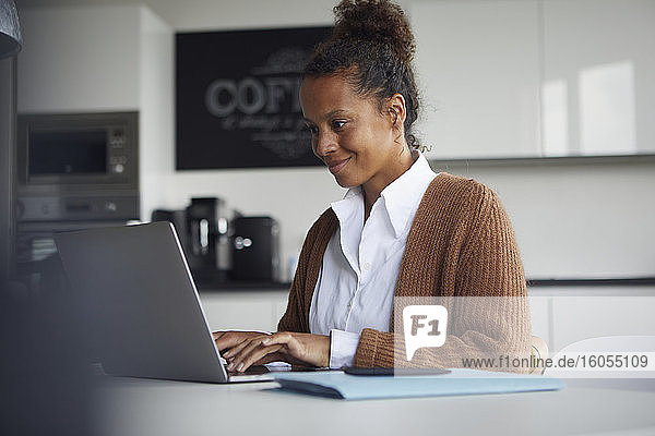 Lächelnde Geschäftsfrau  die am Tisch in der Küche sitzt und einen Laptop benutzt