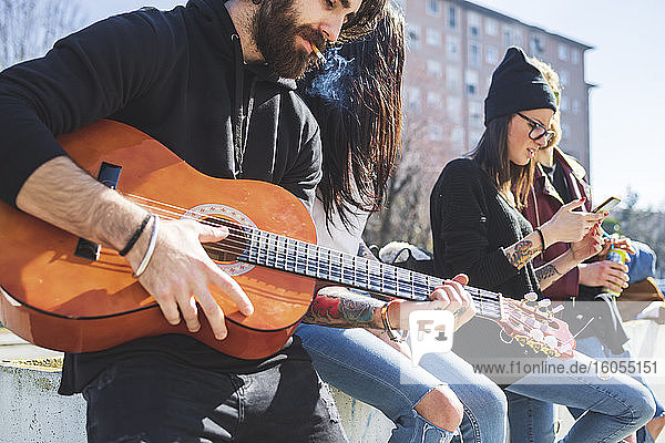 Mann raucht Zigarette und spielt Gitarre  während er mit Freunden in der Stadt chillt