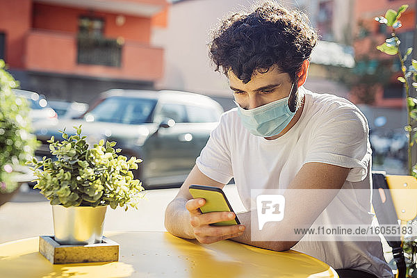 Junger Mann mit Gesichtsmaske  der ein Smartphone benutzt  während er in einem Straßencafé sitzt