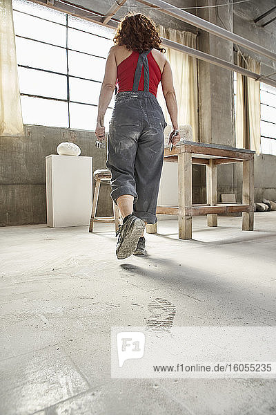 Woman wearing overalls walking on dusty floor towards table in workshop
