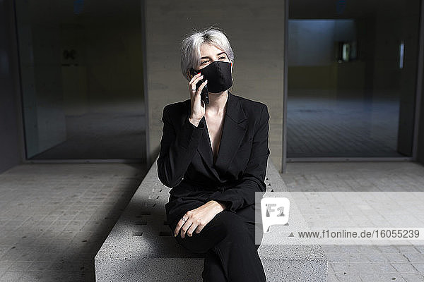 Female professional wearing mask talking over smart phone while sitting in office