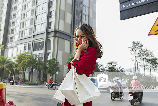 Lächelnde Frau  die Einkaufstaschen trägt und über ihr Smartphone spricht  während sie auf der Straße steht
