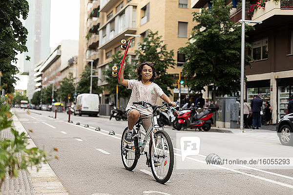 Smiling boy holding skateboard while riding bicycle on street